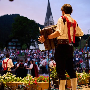 Musiker auf einer Bühne beim Alpenzauber-Festival im Kleinwalsertal, spielt Gitarre während die Menge im Hintergrund zuschaut, mit Bergkulisse und Kirche im Hintergrund. | © Kleinwalsertal Tourismus eGen • Fotograf: Oliver Farys