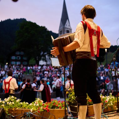 Musiker auf einer Bühne beim Alpenzauber-Festival im Kleinwalsertal, spielt Gitarre während die Menge im Hintergrund zuschaut, mit Bergkulisse und Kirche im Hintergrund. | © Kleinwalsertal Tourismus eGen • Fotograf: Oliver Farys