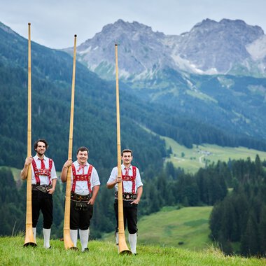 Drei Männer in traditionellen Trachten stehen mit Alphörnern auf einer Wiese vor einer Bergkulisse im Kleinwalsertal. | © Kleinwalsertal Tourismus eGen • Fotograf: Oliver Farys