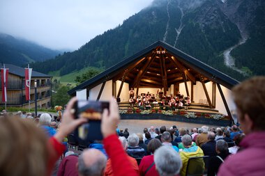 Öffentliches Konzert auf einer offenen Bühne in einem Bergort mit vielen Zuschauern, die das Sommer-Open-Air genießen. | © Kleinwalsertal Tourismus eGen • Fotograf: Oliver Farys