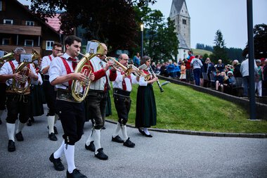 Blasmusikkapelle in traditioneller Tracht beim Umzug in Mittelberg während des Sommerfestivals, mit Publikum im Hintergrund und einem historischen Kirchturm. | © Kleinwalsertal Tourismus eGen • Fotograf: Oliver Farys