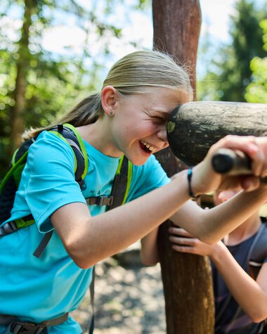 Lächelnde junge Mädchen beim Spielen im Wald, stößt mit einem Holzstück am Kopf an. | © Kleinwalsertal Tourismus eGen • Fotograf: Oliver Farys