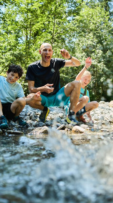 Eine lebhafte Gruppe von vier Menschen, darunter Kinder und ein Erwachsener, sitzt am Ufer eines Baches im Grünen und hat Spaß beim Spielen im Wasser. | © Kleinwalsertal Tourismus eGen • Fotograf: Oliver Farys
