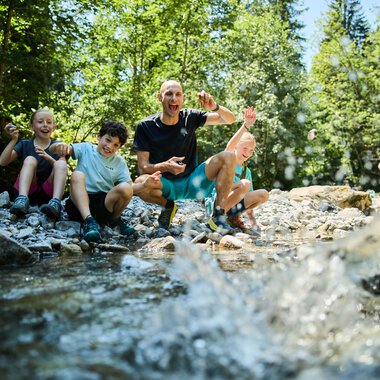 Eine lebhafte Gruppe von vier Menschen, darunter Kinder und ein Erwachsener, sitzt am Ufer eines Baches im Grünen und hat Spaß beim Spielen im Wasser. | © Kleinwalsertal Tourismus eGen • Fotograf: Oliver Farys