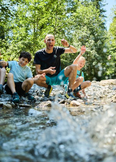 Eine lebhafte Gruppe von vier Menschen, darunter Kinder und ein Erwachsener, sitzt am Ufer eines Baches im Grünen und hat Spaß beim Spielen im Wasser. | © Kleinwalsertal Tourismus eGen • Fotograf: Oliver Farys