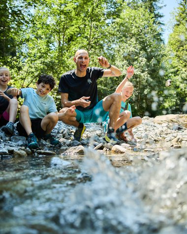 Eine lebhafte Gruppe von vier Menschen, darunter Kinder und ein Erwachsener, sitzt am Ufer eines Baches im Grünen und hat Spaß beim Spielen im Wasser. | © Kleinwalsertal Tourismus eGen • Fotograf: Oliver Farys