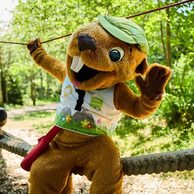 Ein faires Maskottchen in einem Wald bei einer Veranstaltung im Sommer 2024, mit grünem Hut und bunt bedruckter Weste, das auf einem Seil balanciert. | © Kleinwalsertal Tourismus eGen • Fotograf: Oliver Farys