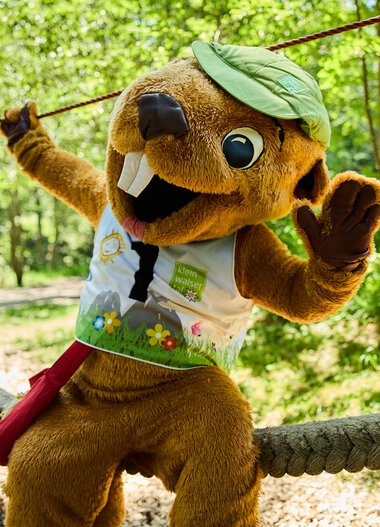 Ein faires Maskottchen in einem Wald bei einer Veranstaltung im Sommer 2024, mit grünem Hut und bunt bedruckter Weste, das auf einem Seil balanciert. | © Kleinwalsertal Tourismus eGen • Fotograf: Oliver Farys