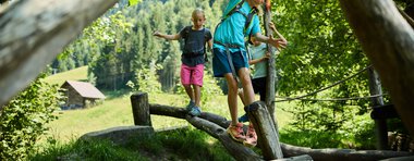 Drei Kinder erkunden einen grünen Waldpfad mit Holzbrücke und bunten Bäumen im Hintergrund, bei schönem Wetter. | © Kleinwalsertal Tourismus eGen • Fotograf: Oliver Farys