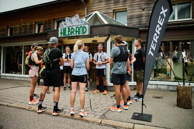Gruppe von Läufern draußen vor einem Laden mit dem Schild 'Hilbrand', die an einem Gespräch teilnehmen, mit einem Adidas Terrex Flag vor der Tür. | © Kleinwalsertal Tourismus eGen • Fotograf: Oliver Farys