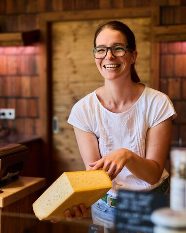 Fröhliche Verkäuferin mit Brille hält ein Stück Käse an einem Hofladen im Kleinwalsertal, im Hintergrund Holzregale und Verkaufsartikel. | © Kleinwalsertal Tourismus eGen • Fotograf: Oliver Farys