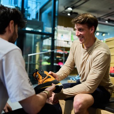 Zwei Männer handeln in einem Sportgeschäft mit Wanderschuhen. Einer übergibt dem anderen einen Schuh, beide lachen. | © Kleinwalsertal Tourismus eGen • Fotograf: Oliver Farys
