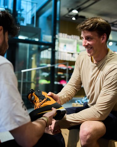Zwei Männer handeln in einem Sportgeschäft mit Wanderschuhen. Einer übergibt dem anderen einen Schuh, beide lachen. | © Kleinwalsertal Tourismus eGen • Fotograf: Oliver Farys