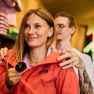 Junge Frau beim Einkauf in einem Geschäft, trägt eine orangefarbene Jacke und hält einen Löffel in der Hand. | © Kleinwalsertal Tourismus eGen • Fotograf: Oliver Farys