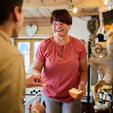 Freundliche Verkäuferin in einem Laden, die mit einem Kunden spricht, während sie eine Scheibe Käse hält. | © Kleinwalsertal Tourismus eGen • Fotograf: Oliver Farys