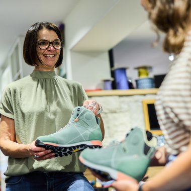 Frau mit Brille und kurzen Haaren hält ein Paar blaue Wanderschuhe und spricht mit einer Kundin im Laden. | © Kleinwalsertal Tourismus eGen • Fotograf: Oliver Farys