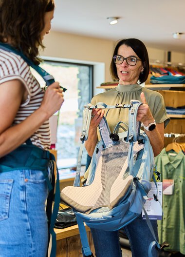 Zwei Frauen in einem Outdoor-Geschäft, eine zeigt der anderen einen gepolsterten Rucksack. Im Hintergrund sind Kleiderregale mit bunten Jacken und Outdoor-Ausrüstung. | © Kleinwalsertal Tourismus eGen • Fotograf: Oliver Farys
