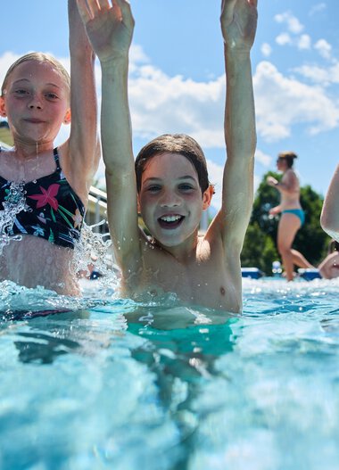 Drei Kinder erfreuen sich im Freibad, lachen und haben Spaß im Wasser, mit einem blauen Himmel und Sonnenlicht im Hintergrund. | © Kleinwalsertal Tourismus eGen • Fotograf: Oliver Farys