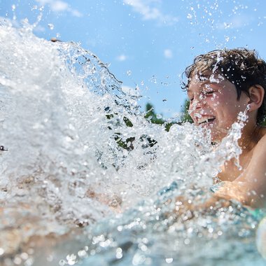 Eine fröhliche Frau plantscht im Freibad bei strahlendem Sonnenschein, Wasser spritzt um sie herum. | © Kleinwalsertal Tourismus eGen • Fotograf: Oliver Farys