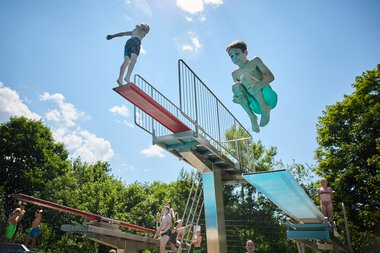 Kinder springen von einem Wasser-Sprungbrett im Freibad Riezlern, umgeben von üppigem Grün bei sonnigem Himmel. | © Kleinwalsertal Tourismus eGen • Fotograf: Oliver Farys