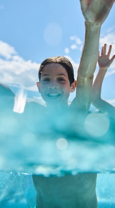 Zwei Kinder im Freibad Riezlern, lachen und planschen im Wasser bei Sonnenschein, mit blauen Himmel und Wolken im Hintergrund. | © Kleinwalsertal Tourismus eGen • Fotograf: Oliver Farys
