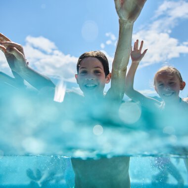 Zwei Kinder im Freibad Riezlern, lachen und planschen im Wasser bei Sonnenschein, mit blauen Himmel und Wolken im Hintergrund. | © Kleinwalsertal Tourismus eGen • Fotograf: Oliver Farys