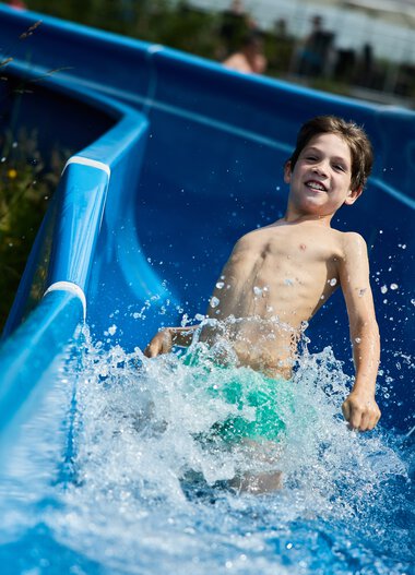 Junge, der fröhlich auf einer Wasserrutschbahn im Freibad Riezlern im Kleinwalsertal Spaß hat. | © Kleinwalsertal Tourismus eGen • Fotograf: Oliver Farys