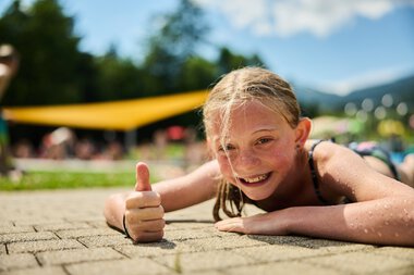 Ein lachendes Mädchen liegt am Boden eines Freibads in Riezlern, zeigt mit dem Daumen nach oben bei strahlendem Sommerwetter. | © Kleinwalsertal Tourismus eGen • Fotograf: Oliver Farys