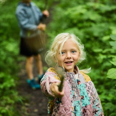 Zwei Kinder erkunden einen Wald, das Mädchen im Vordergrund freut sich und zeigt nach vorne, während das andere Kind im Hintergrund abseits steht, umgeben von grünen Bäumen. | © Kleinwalsertal Tourismus eGen • Fotograf: Oliver Farys