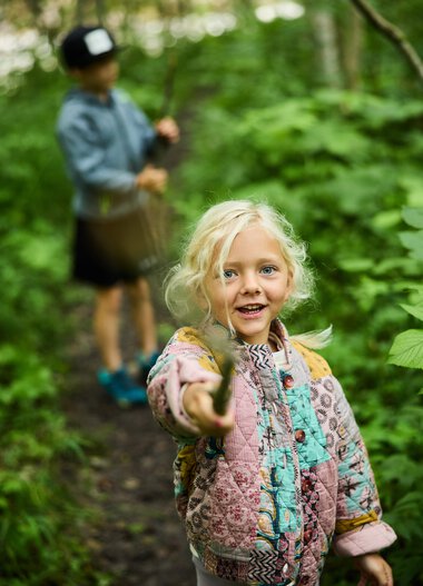 Zwei Kinder erkunden einen Wald, das Mädchen im Vordergrund freut sich und zeigt nach vorne, während das andere Kind im Hintergrund abseits steht, umgeben von grünen Bäumen. | © Kleinwalsertal Tourismus eGen • Fotograf: Oliver Farys