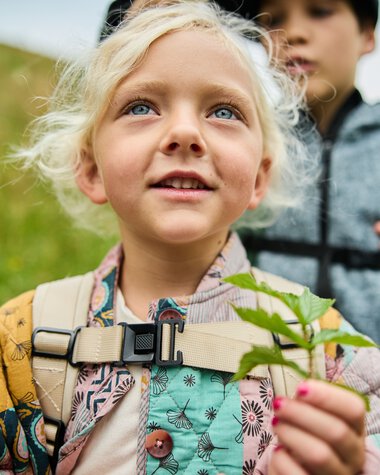 Kinder beim Naturerlebnis im Sommer 2024, ein Mädchen mit Rucksack hält eine Pflanze, umgeben von grüner Landschaft. | © Kleinwalsertal Tourismus eGen • Fotograf: Oliver Farys