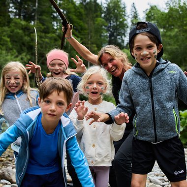 Fünf fröhliche Kinder beim Sommerabenteuer im Freien, lächelnd und spielend an einem Fluss, umgeben von grüner Natur. | © Kleinwalsertal Tourismus eGen • Fotograf: Oliver Farys