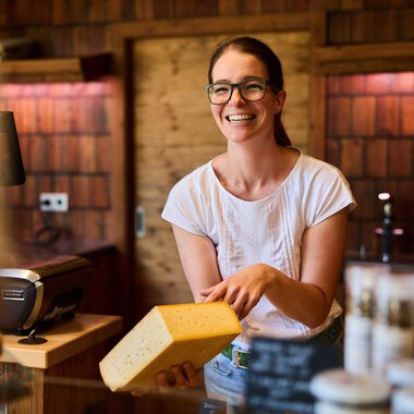 Smiling female vendor with glasses holding a piece of cheese at a farm shop in Kleinwalsertal, with wooden shelves and products in the background. | © Kleinwalsertal Tourismus eGen • Photographer: Oliver Farys