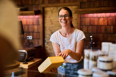Smiling female vendor with glasses holding a piece of cheese at a farm shop in Kleinwalsertal, with wooden shelves and products in the background. | © Kleinwalsertal Tourismus eGen • Photographer: Oliver Farys
