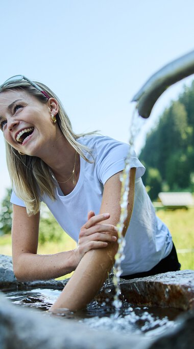 Frau sitzt am Kneippbecken, lacht und nutzt die kalte Wassertherapie im Grünen bei schönem Wetter. | © Kleinwalsertal Tourismus eGen • Fotograf: Oliver Farys
