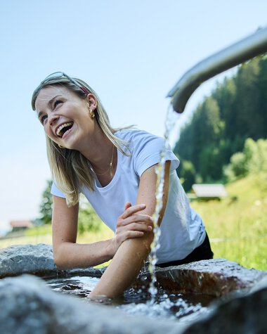 Frau sitzt am Kneippbecken, lacht und nutzt die kalte Wassertherapie im Grünen bei schönem Wetter. | © Kleinwalsertal Tourismus eGen • Fotograf: Oliver Farys