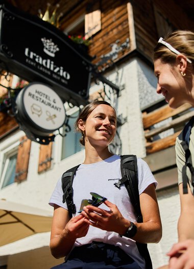 Zwei junge Frauen mit Rucksäcken sprechen lachend vor einem Bergrestaurant in Kleinwalsertal bei sonnigem Wetter. | © Kleinwalsertal Tourismus eGen • Fotograf: Oliver Farys