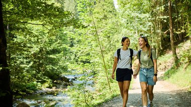 © Kleinwalsertal Tourismus eGen • Fotograf: Oliver Farys Zwei junge Frauen wandern lachend durch einen grünen Waldweg am Fluss im Sommer 2024, begleitet von Natur und Sonnenschein. | © Kleinwalsertal Tourismus eGen • Fotograf: Oliver Farys