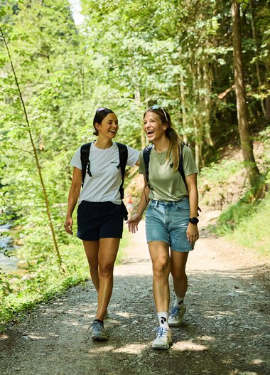 Zwei junge Frauen wandern lachend durch einen grünen Waldweg am Fluss im Sommer 2024, begleitet von Natur und Sonnenschein. | © Kleinwalsertal Tourismus eGen • Fotograf: Oliver Farys
