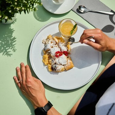 Table with a plate of delicious Kaiserschmarrn dusted with powdered sugar and garnished with cherries, alongside a glass of fresh orange juice. | © Kleinwalsertal Tourismus eGen • Photographer: Oliver Farys