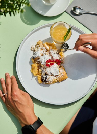 Tisch mit einem Teller mit köstlichem Kaiserschmarrn, garnierte mit Puderzucker und Kirschen, daneben ein Glas frisch gepresster Orangensaft. | © Kleinwalsertal Tourismus eGen • Fotograf: Oliver Farys