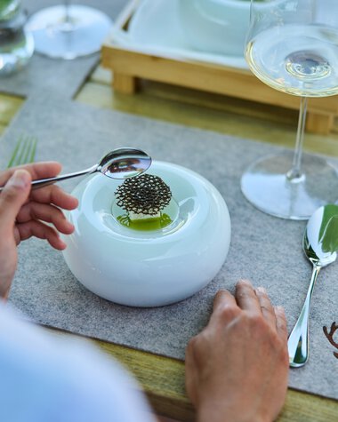 Ein gedeckter Tisch mit einer modernen weißen Platte, auf der ein Teller mit einem kleinen Gericht und Stäbchen liegt. Es gibt auch ein Glas Weißwein, ein Wasserglas mit Minzblättern und eine silberne Gabel. Der Tisch hat eine Holzoptik und eine braune Decke mit Jagdmotiv. | © Kleinwalsertal Tourismus eGen • Fotograf: Oliver Farys
