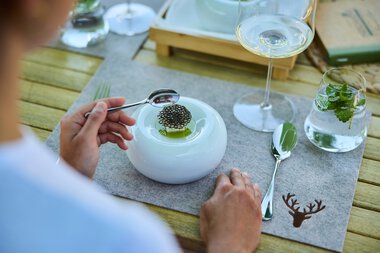 Ein gedeckter Tisch mit einer modernen weißen Platte, auf der ein Teller mit einem kleinen Gericht und Stäbchen liegt. Es gibt auch ein Glas Weißwein, ein Wasserglas mit Minzblättern und eine silberne Gabel. Der Tisch hat eine Holzoptik und eine braune Decke mit Jagdmotiv. | © Kleinwalsertal Tourismus eGen • Fotograf: Oliver Farys