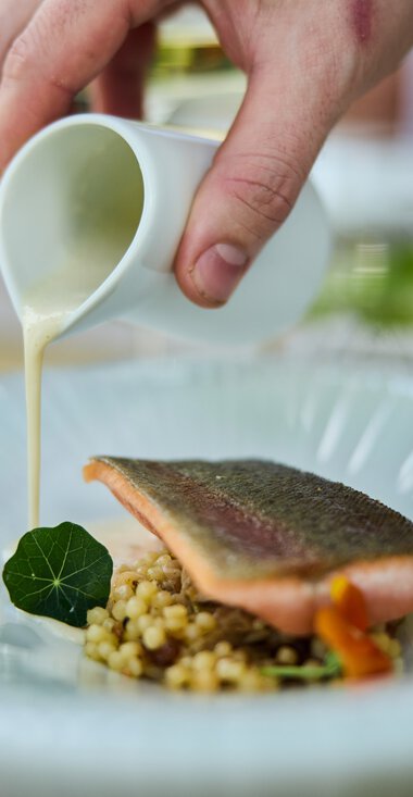 Ein Koch gießt cremige Sauce über ein Gericht mit Fisch, serviert auf einem weißen Teller bei einem kulinarischen Event. | © Kleinwalsertal Tourismus eGen • Fotograf: Oliver Farys