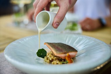 Ein Koch gießt cremige Sauce über ein Gericht mit Fisch, serviert auf einem weißen Teller bei einem kulinarischen Event. | © Kleinwalsertal Tourismus eGen • Fotograf: Oliver Farys
