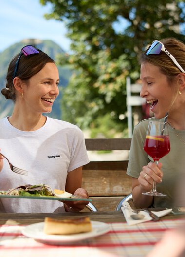 Zwei lachende Frauen genießen zusammen ein Peak-Lunch im Freien mit Bergpanorama im Hintergrund, bei sommerlichem Wetter. | © Kleinwalsertal Tourismus eGen • Fotograf: Oliver Farys