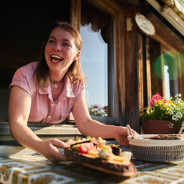 A smiling young woman stands at a window of a rustic mountain hut, holding a served meal and enjoying the summer atmosphere in Kleinwalsertal. | © Kleinwalsertal Tourismus eGen • Photographer: Oliver Farys