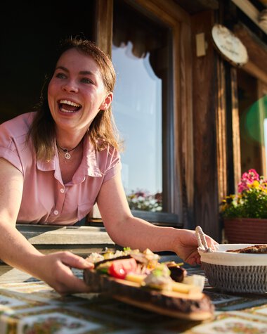Eine lachende junge Frau steht an einem Fenster der almähnlichen Hütte, hält eine servierte Mahlzeit und genießt die Sommeratmosphäre im Kleinwalsertal. | © Kleinwalsertal Tourismus eGen • Fotograf: Oliver Farys