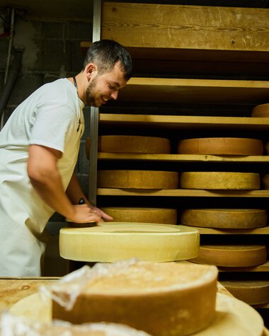 Mann in Weißkochkleid arbeitet in einer Käsekeller- oder Käseherstellungsumgebung, umgeben von runden Käse reifen. | © Kleinwalsertal Tourismus eGen • Fotograf: Oliver Farys