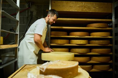 Mann in Weißkochkleid arbeitet in einer Käsekeller- oder Käseherstellungsumgebung, umgeben von runden Käse reifen. | © Kleinwalsertal Tourismus eGen • Fotograf: Oliver Farys